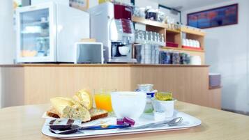 A breakfast tray with bread, juice, and condiments on a table in a cafe with a coffee machine and shelves in the background.