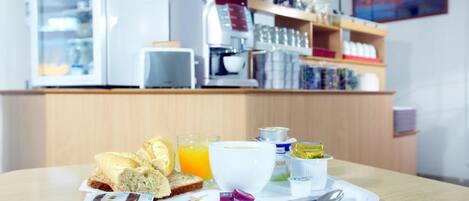 A breakfast tray with bread, juice, and condiments on a table in a cafe with a coffee machine and shelves in the background.