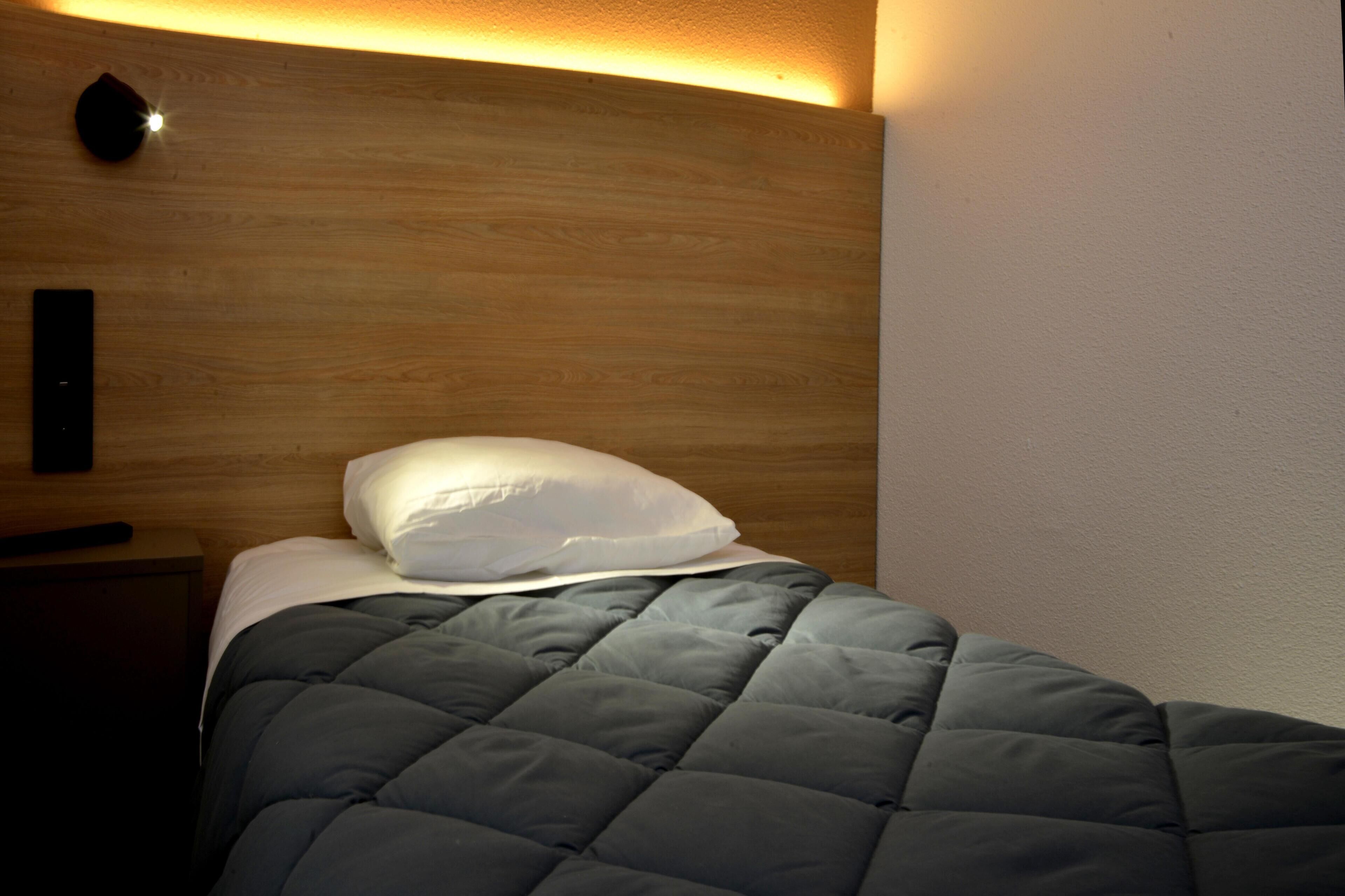 A neatly made bed with a dark quilt and a white pillow, a wooden headboard, and a wall-mounted light fixture.