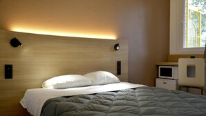 A neatly made bed with a gray comforter and white pillows, a bedside table with a white appliance, and a window with a view of greenery. - Premiere Classe Dunkerque Sud - Loon Plage (Loon-Plage)