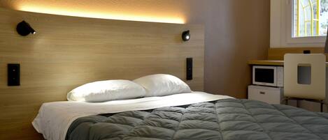 Una cama bien hecha con una colcha gris y almohadas blancas, una mesita de noche con un electrodoméstico blanco, y una ventana con vistas a zonas verdes.