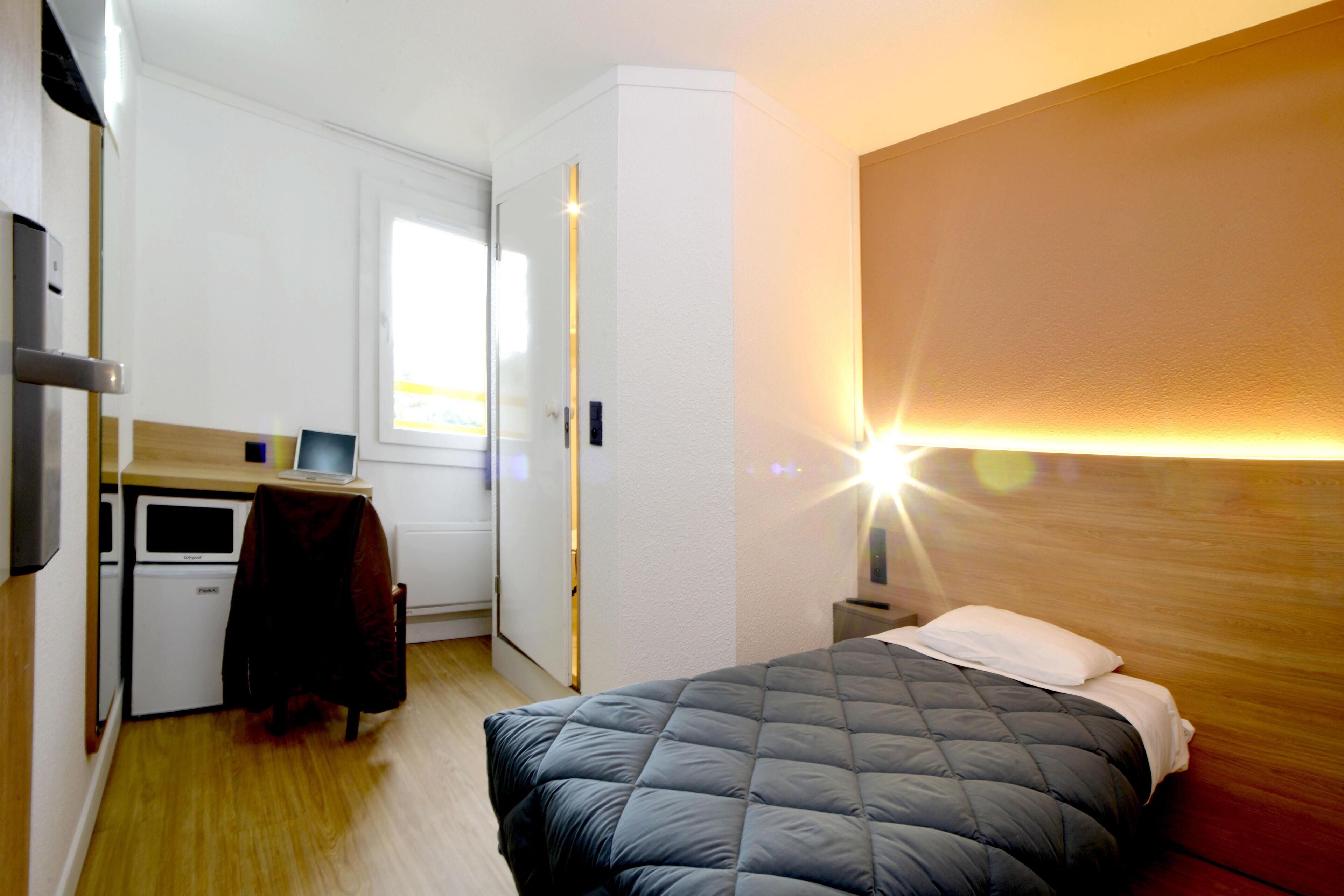 Una habitación de hotel con cama, escritorio, silla, perchero, armario y mesita de noche con lámpara.