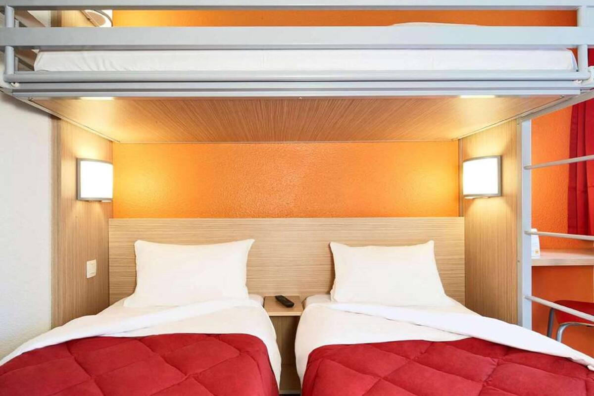 A bunk bed room with two beds, each with a red quilted cover, and a wooden headboard. There are two wall-mounted lights above the beds.