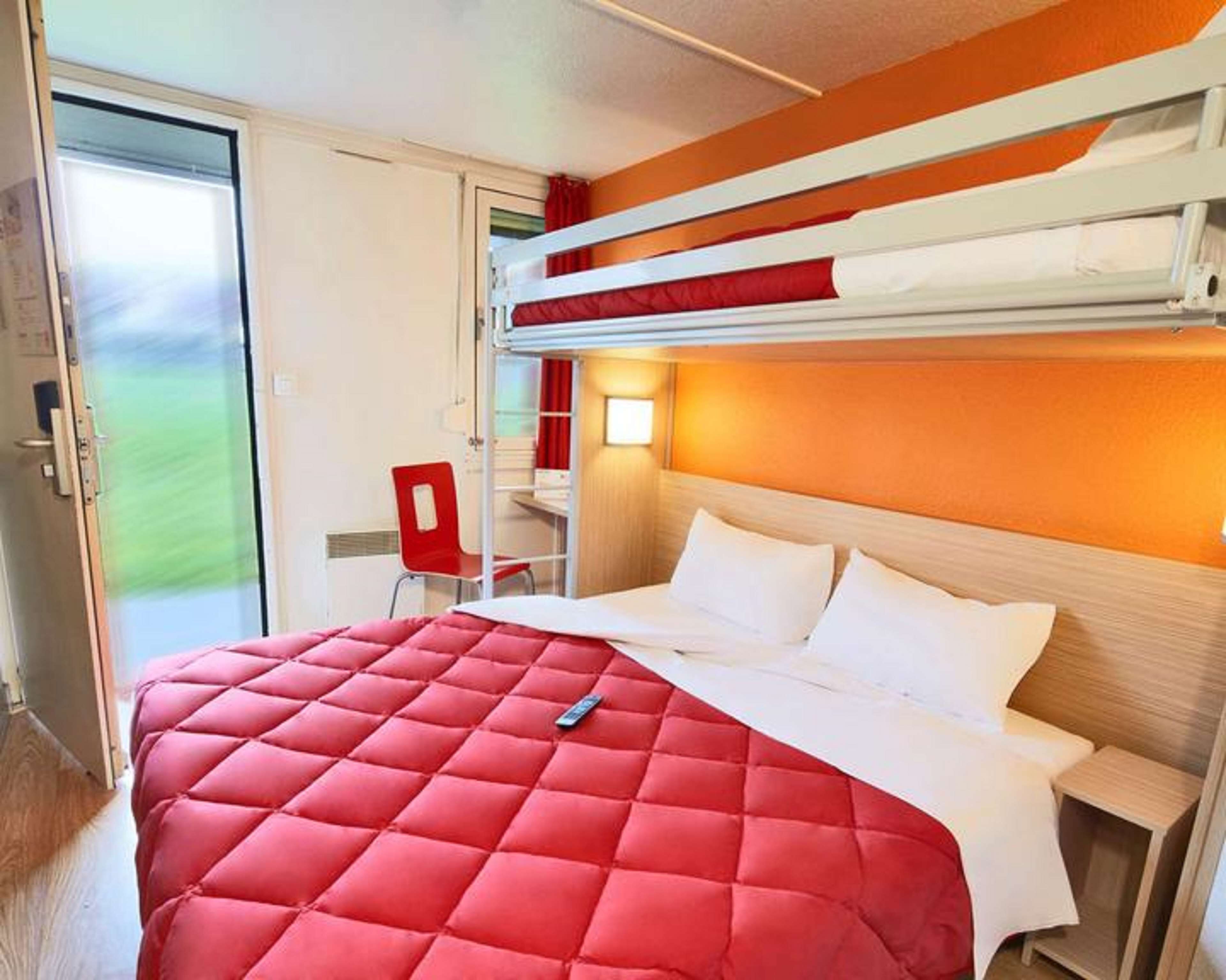 Une chambre d’hôtel avec un lit superposé, une couverture matelassée rouge, un bureau avec une chaise et une fenêtre donnant sur un paysage verdoyant.