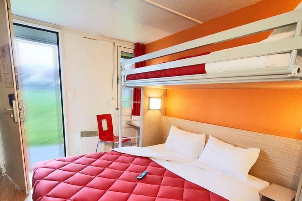 A hotel room with a bunk bed, a red quilted bedspread, a desk with a chair, and a window with a view of greenery.