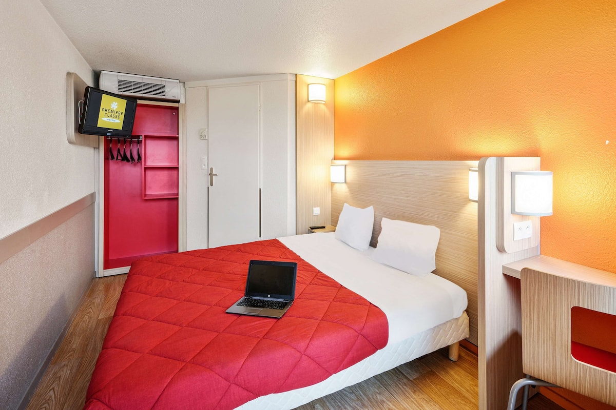 A hotel room with a bed, a laptop on the bed, a red bedspread, a wooden nightstand, a red chair, a white wall, and a TV mounted on the wall.