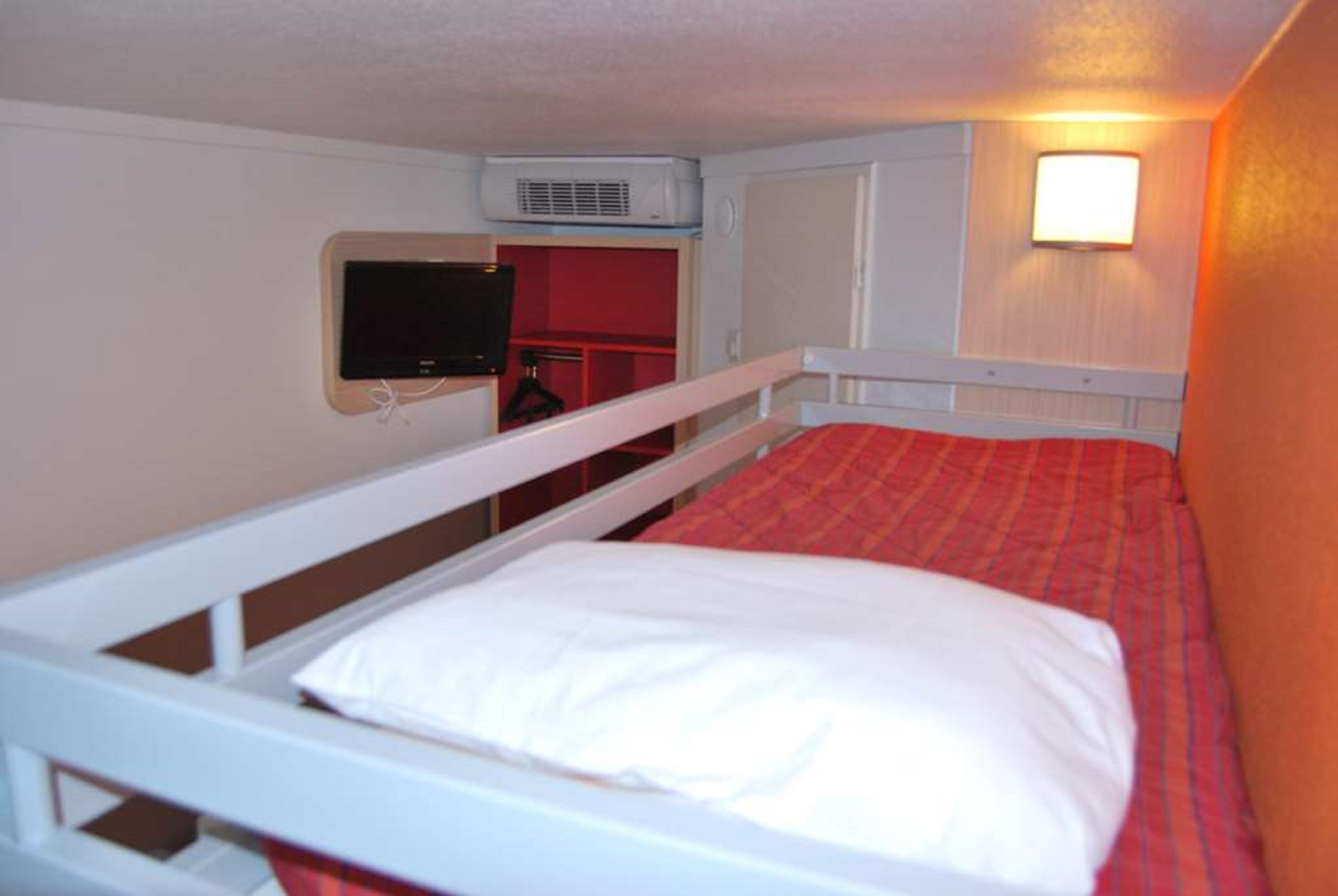 A single-bed room with a red bedspread, white bedding, a mounted television, and a wall-mounted air conditioner.