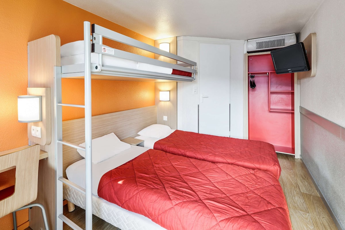 A bunk bed with a red quilted bedspread, a desk with a chair, a wardrobe, and a television mounted on the wall.