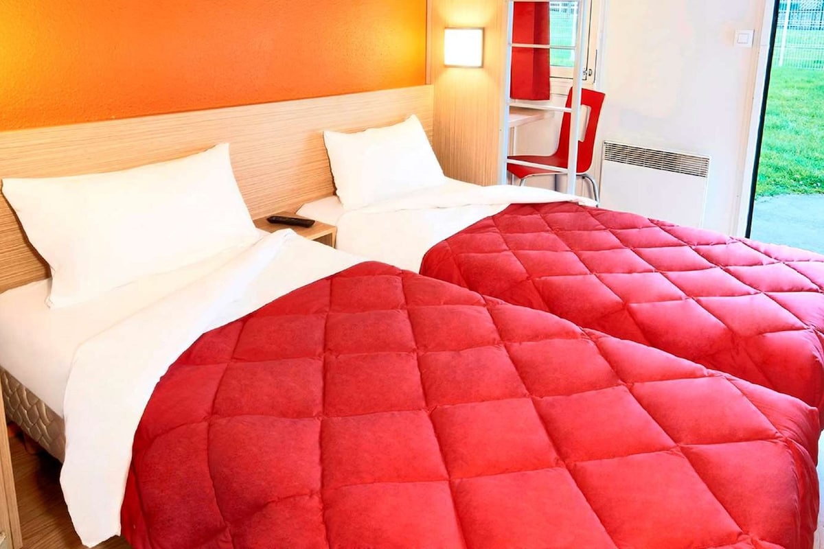 A double bed with a red quilted cover and white pillows in a room with wooden headboard and a window with a view of greenery.