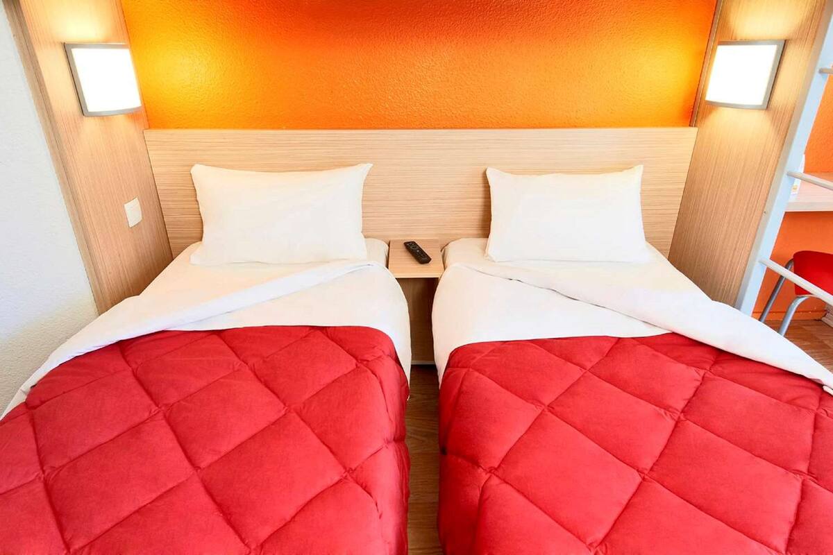 A hotel room with two beds, each with a red quilted bedspread and white pillows. There is a wooden headboard with a warm light above it, and a small table between the beds.