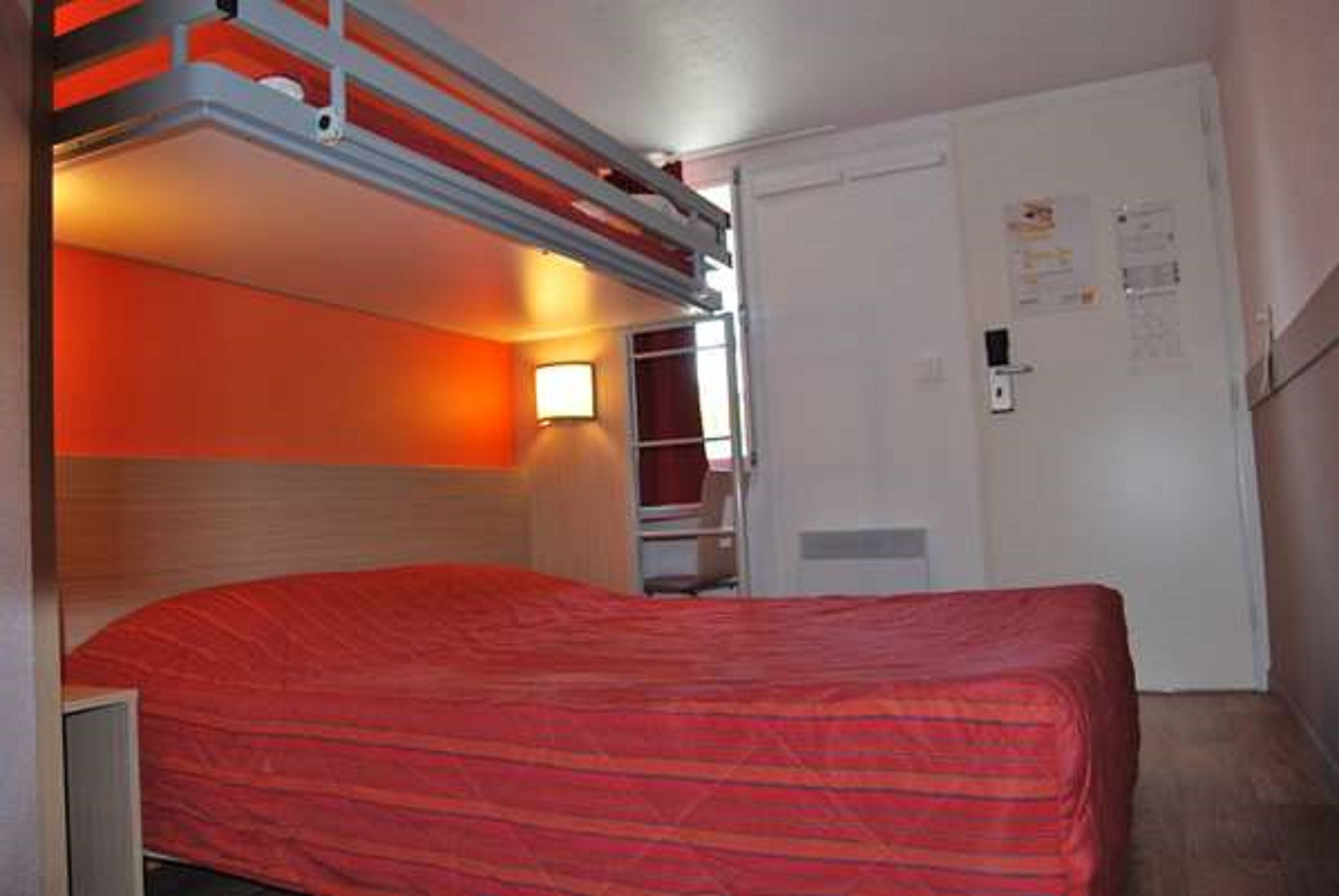 A hotel room with a bunk bed, a red bedspread, a bedside table, and a wall-mounted light.