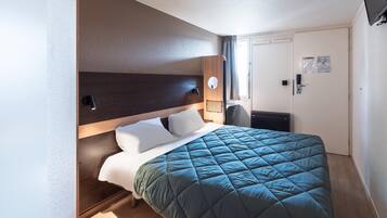 Une chambre d’hôtel avec un lit, une table de chevet, une lampe fixée au mur et une porte dotée d’une poignée.