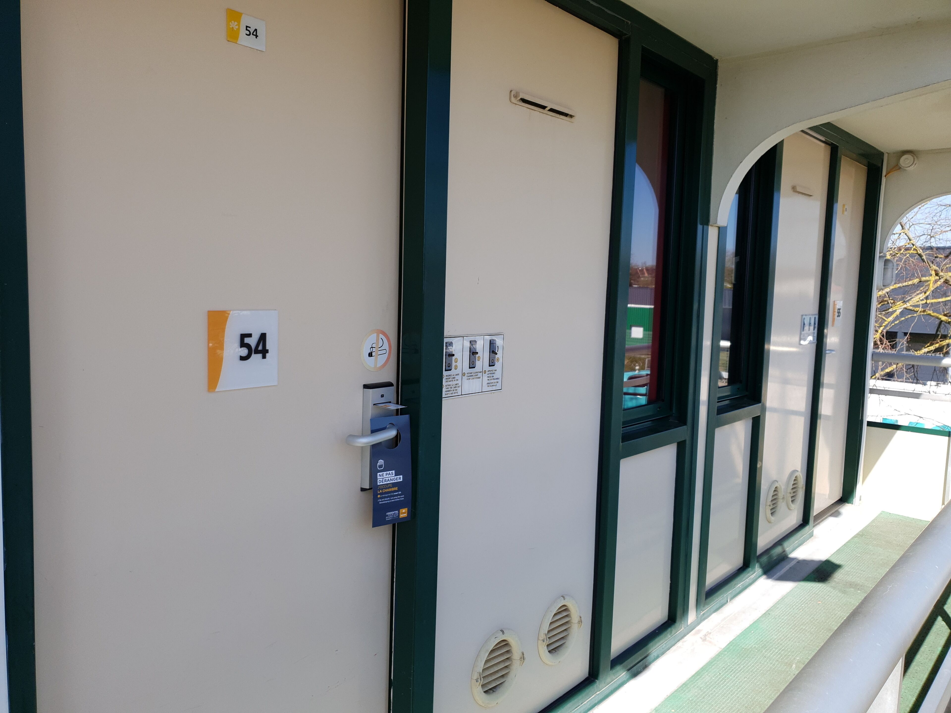 Een hotelkamer met een deur waarop '54' staat, een raam met uitzicht op een gebouw en een deurbelknop.