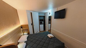 Une chambre d’hôtel de taille réduite, comprenant un lit, une table de chevet, un téléviseur fixé au mur et une armoire avec un miroir.