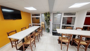 A conference room with white tables and wooden chairs, a large flat-screen TV on the wall, and a potted plant.