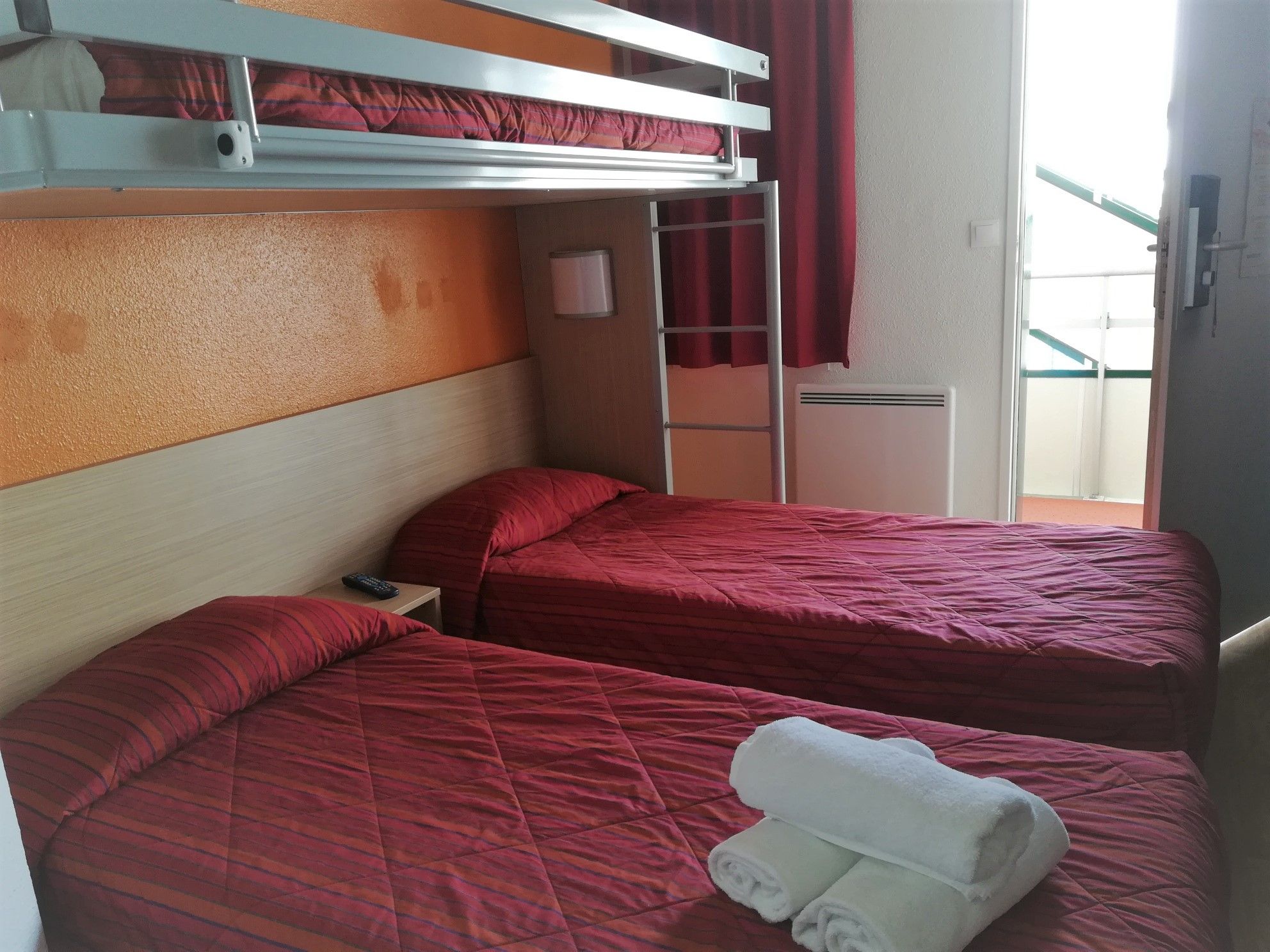 A room with two beds, each with red bedding, a small table with a remote control, and a window with curtains.