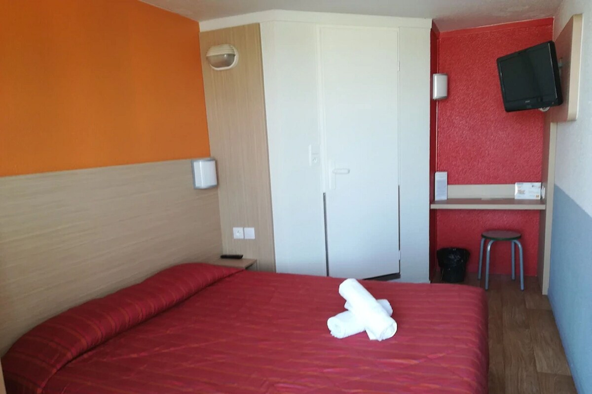 Habitación de hotel con cama roja, puerta blanca, escritorio pequeño con silla y televisor montado en la pared.