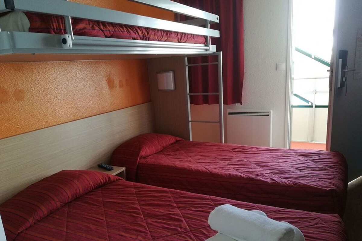Een hotelkamer met twee bedden, een stapelbed bovenop, een klein nachtkastje met een lamp en een raam met gordijnen.