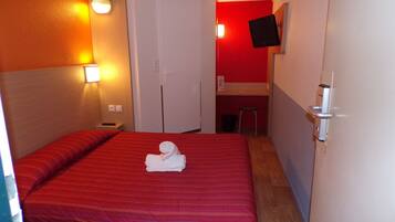 Habitación de hotel con una cama de rayas rojas, una toalla blanca, un escritorio con una silla y un televisor fijado a la pared.