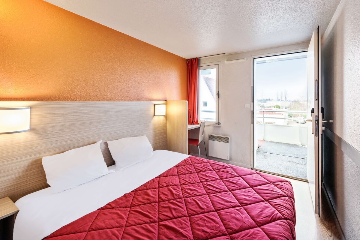 Habitación de hotel con cama, escritorio y vista de la ciudad a través de una puerta corrediza.