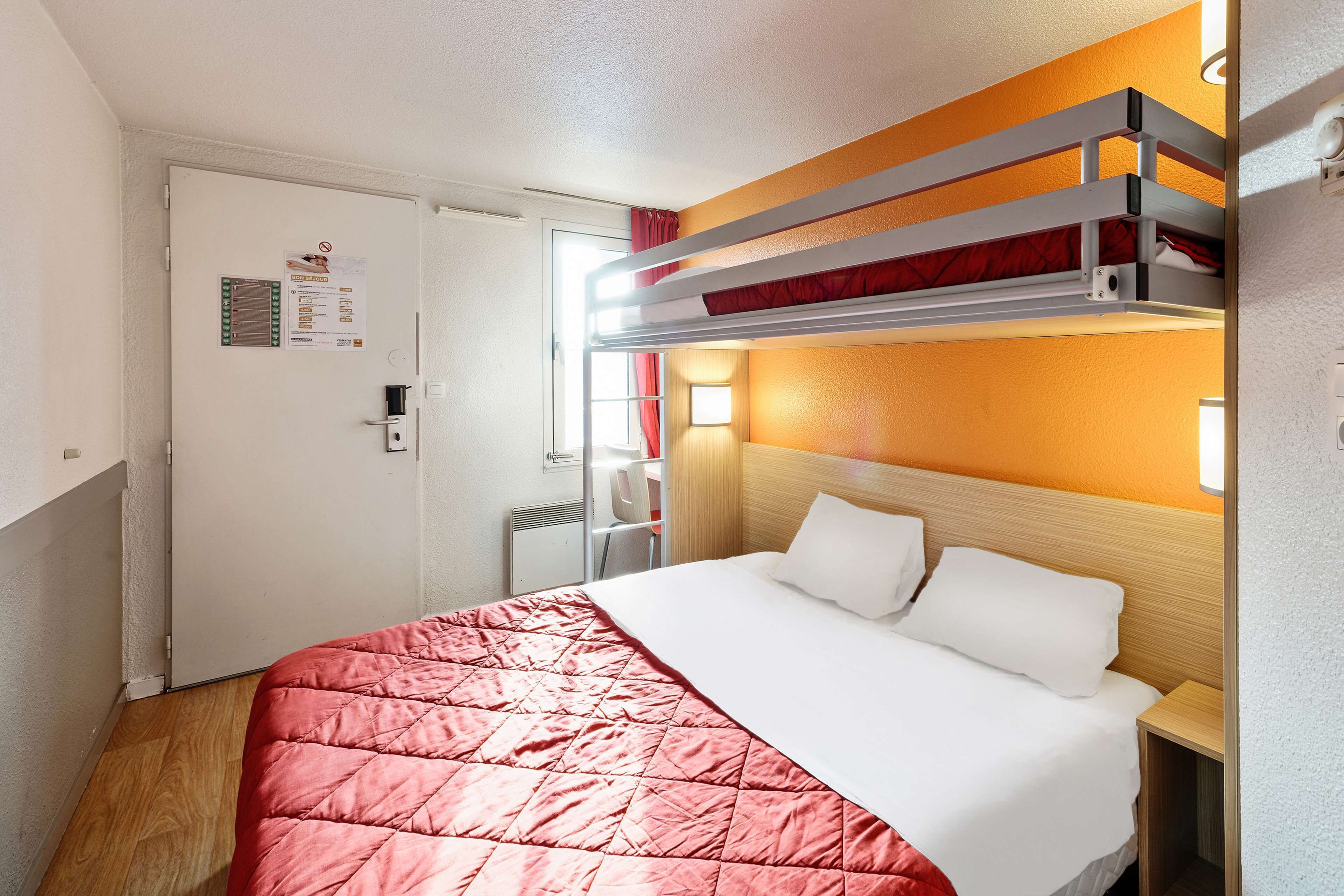A hotel room with a bunk bed, a single bed, a wooden headboard, a red quilted bedspread, a white pillow, a bedside table, and a door with a notice.
