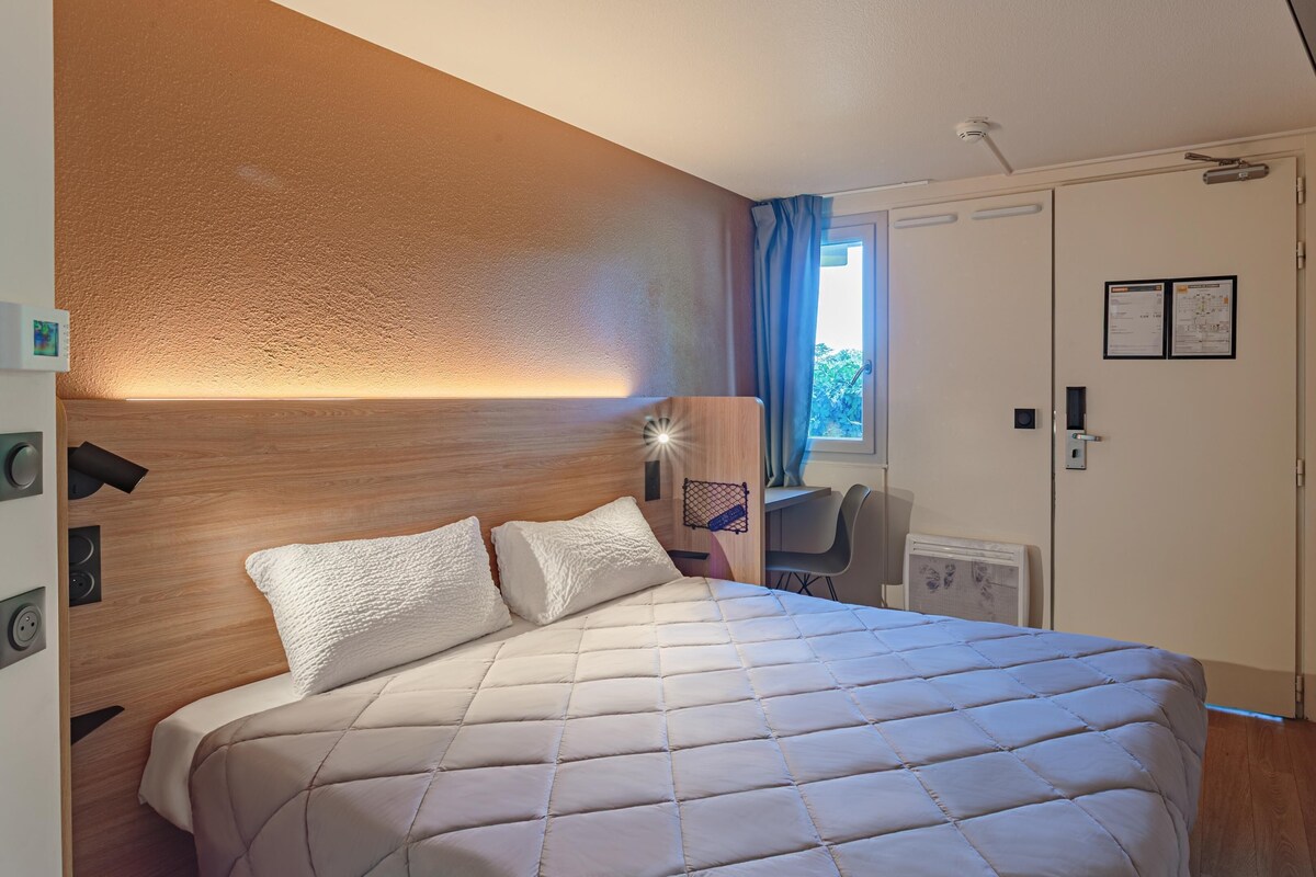 A hotel room with a bed, a desk, a chair, a window with curtains, a door, and a wall-mounted light fixture.