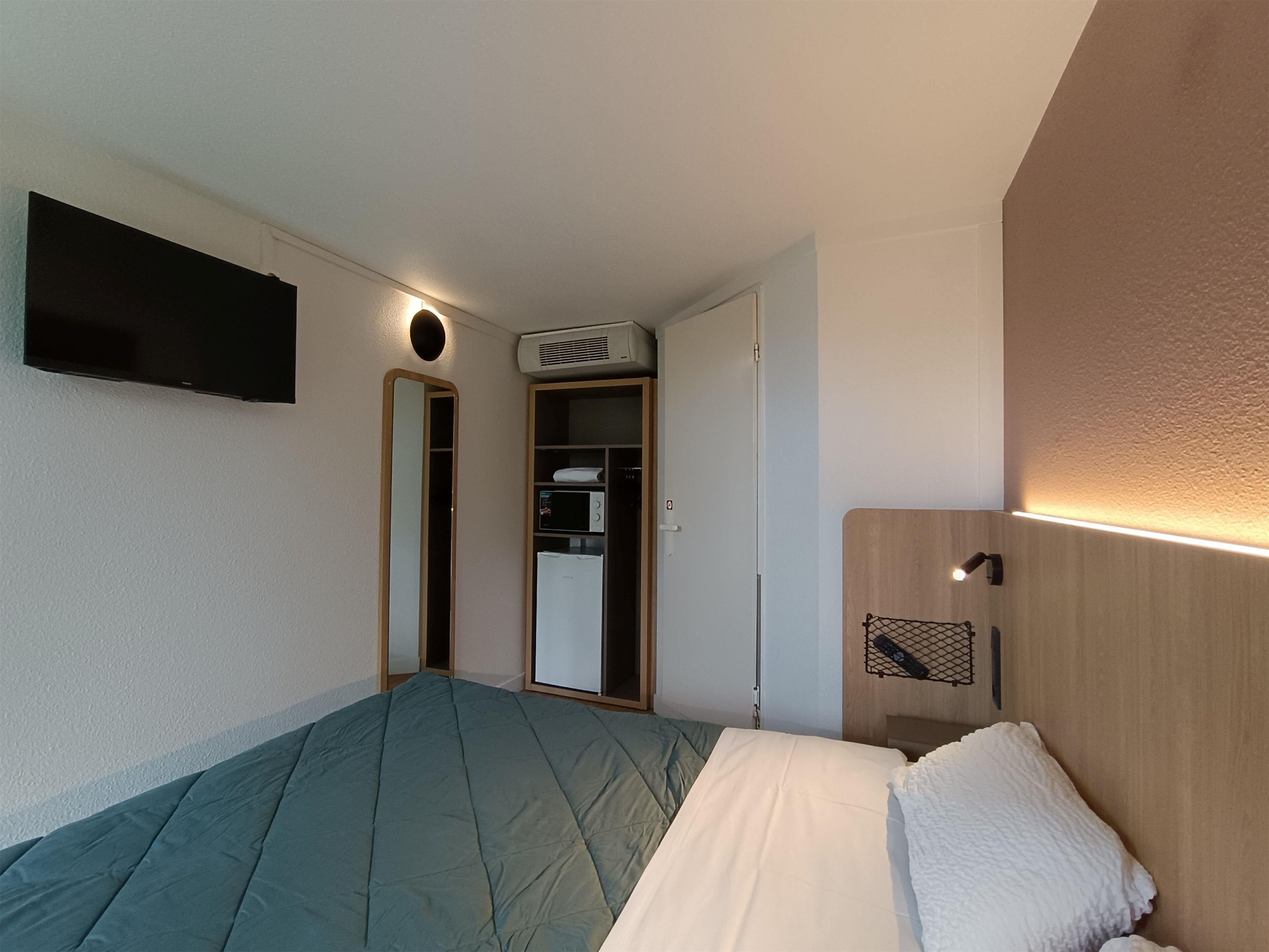 A hotel room with a bed, a flat-screen TV mounted on the wall, a wardrobe, and a small bedside table with a lamp.