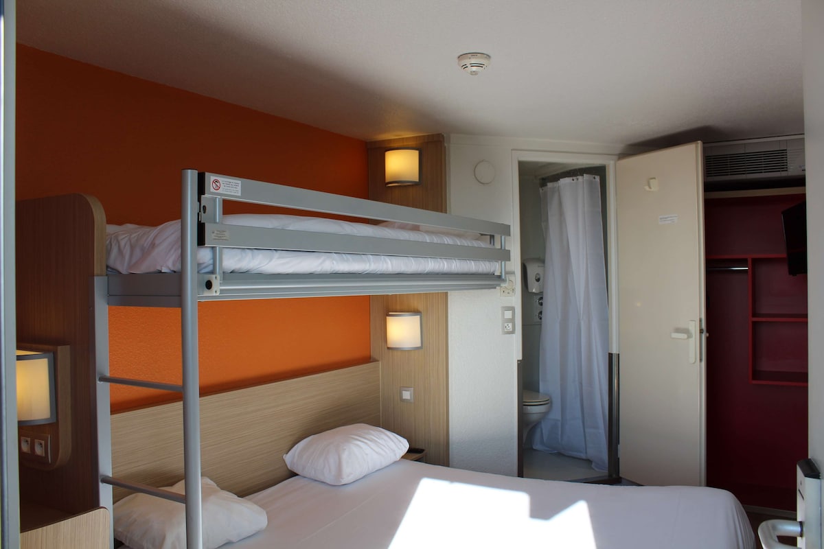 Une chambre avec un lit superposé, une tête de lit en bois, une table de chevet avec une lampe et une porte de salle de bain.