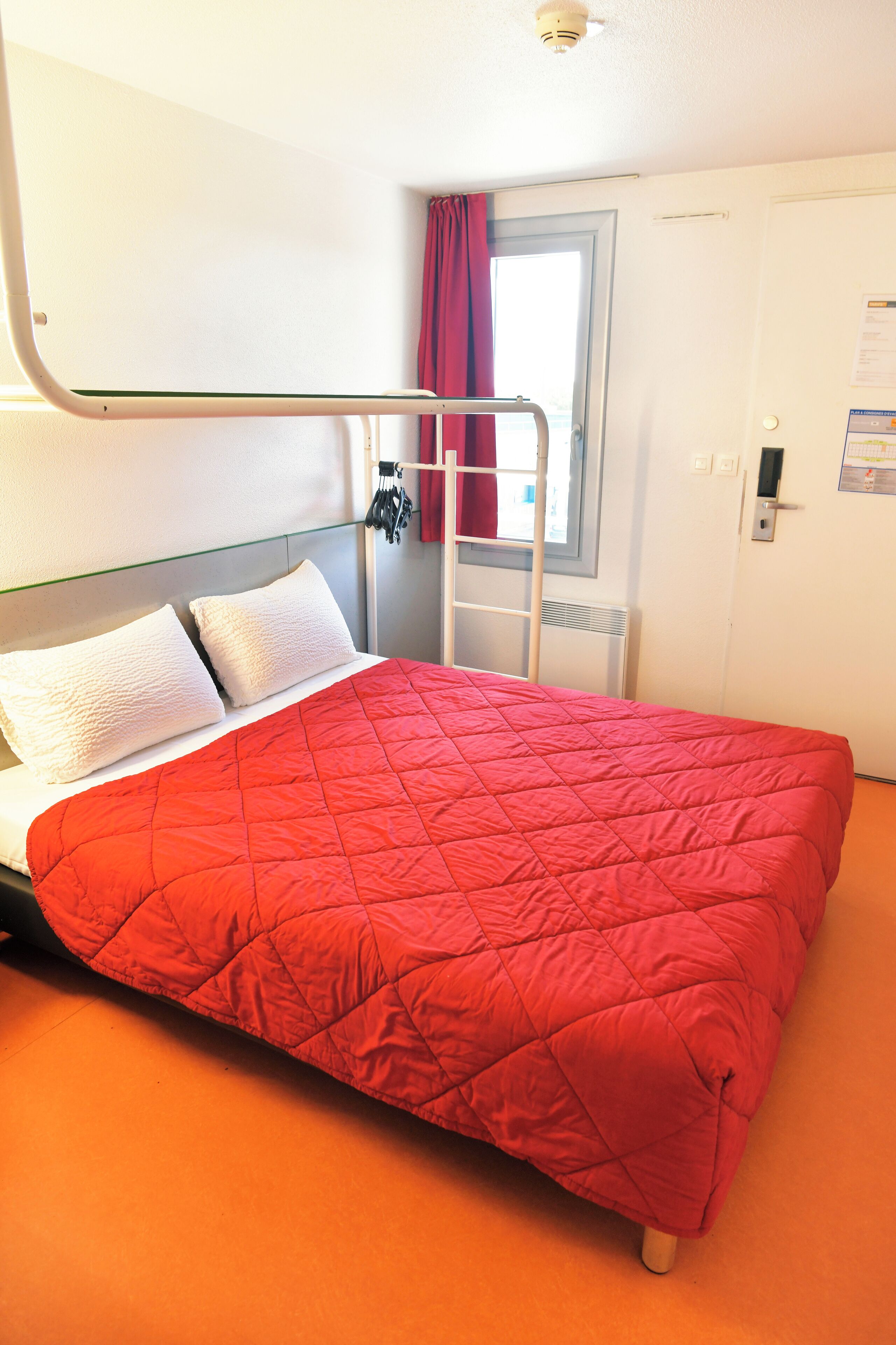 Een kamer met een enkel bed, een rode gewatteerde sprei, witte kussens, een metalen trap, een raam met rode gordijnen en een witte deur met een klein bordje.