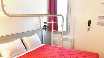Een kamer met een enkel bed, een rode gewatteerde sprei, witte kussens, een metalen trap, een raam met rode gordijnen en een witte deur met een klein bordje.
