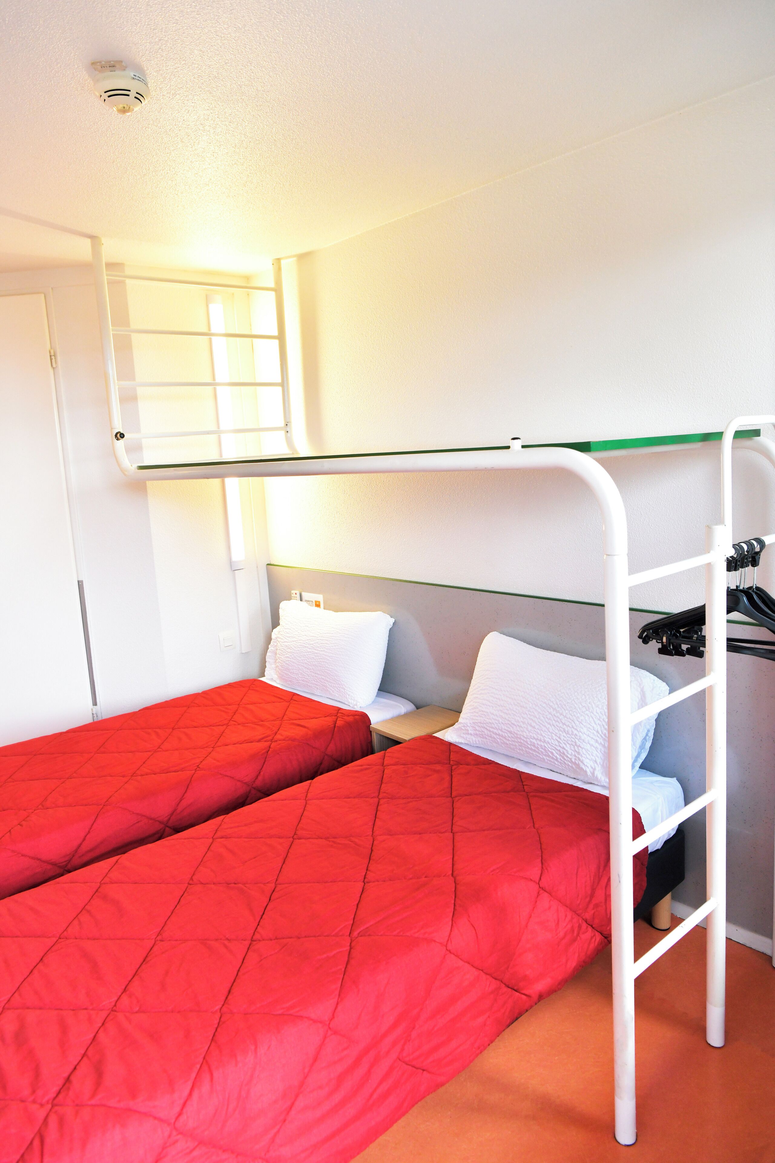 Un letto a castello con una coperta rossa, un materasso bianco e una testiera bianca. Si vede una porta bianca e una finestra con persiane.