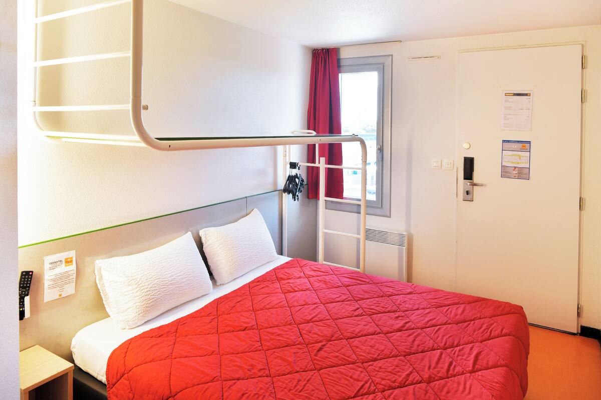 Une chambre d’hôtel avec un lit superposé, une couvre-lit rouge matelassée, un oreiller blanc, une petite table de chevet et une fenêtre avec un rideau rouge.