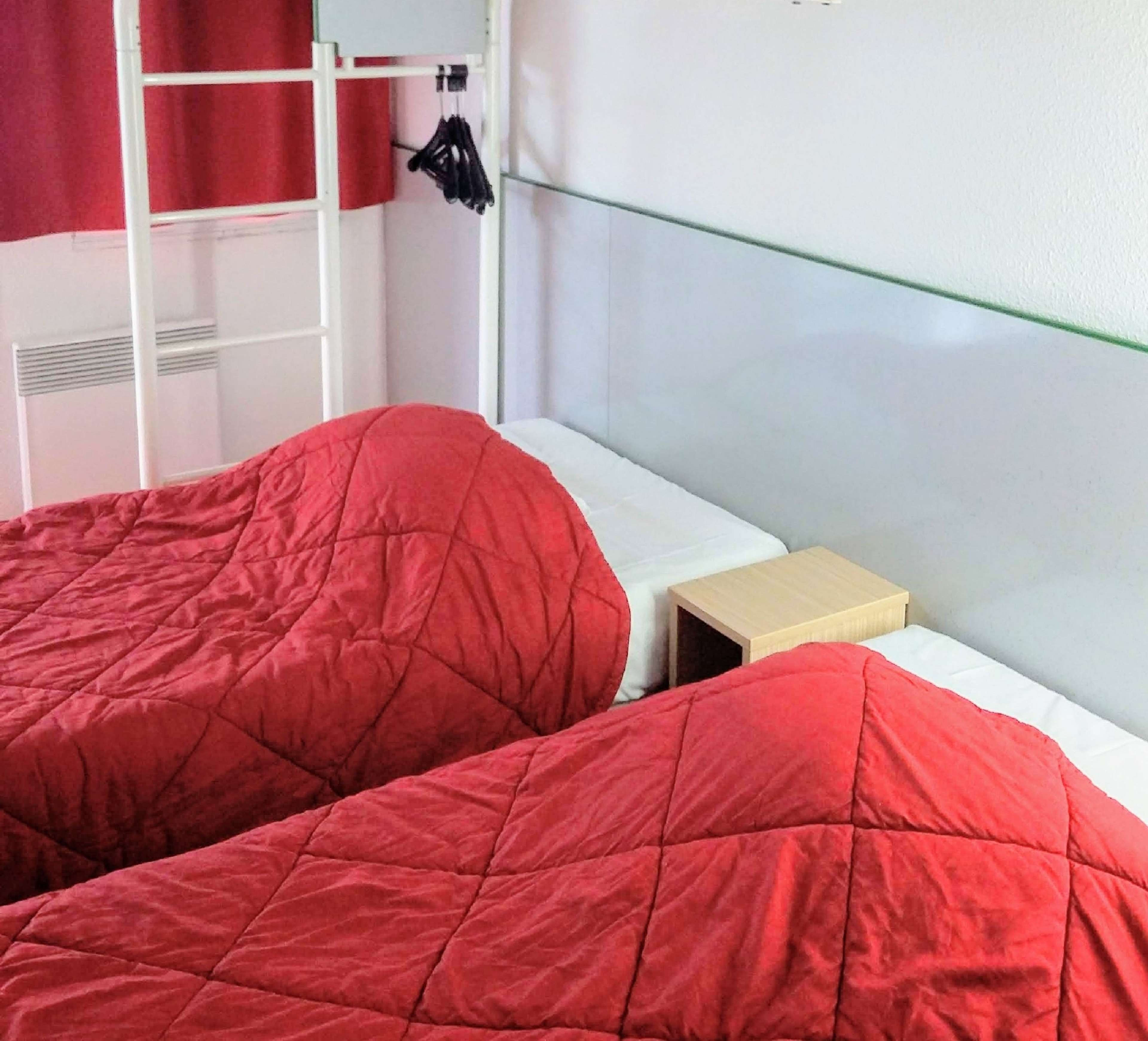 Une chambre avec deux lits, chacun recouvert d’une literie rouge, une petite table de chevet et un mur blanc avec une fenêtre.
