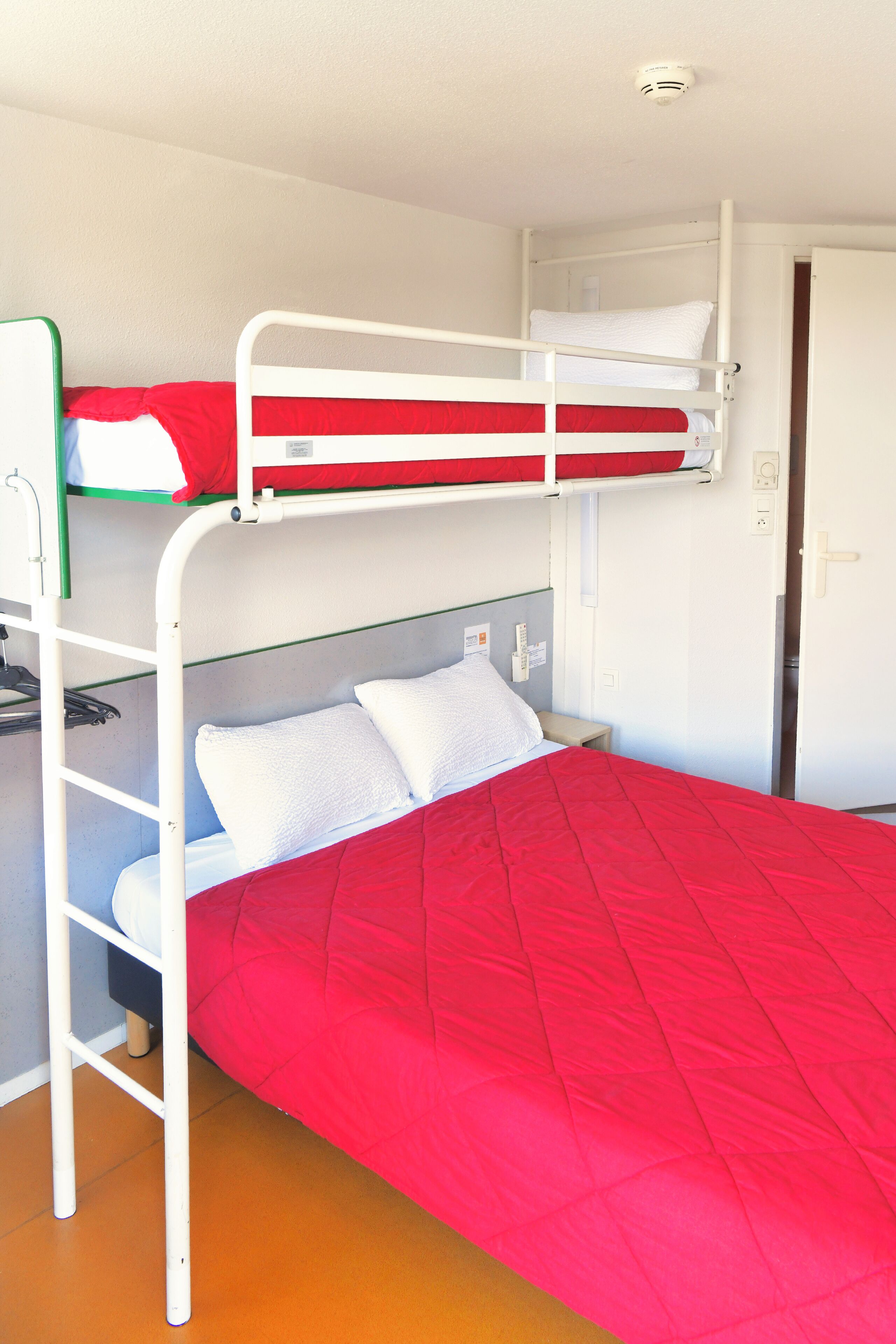 A bunk bed with a red mattress and white pillows, a white door, and a small shelf with a green item.