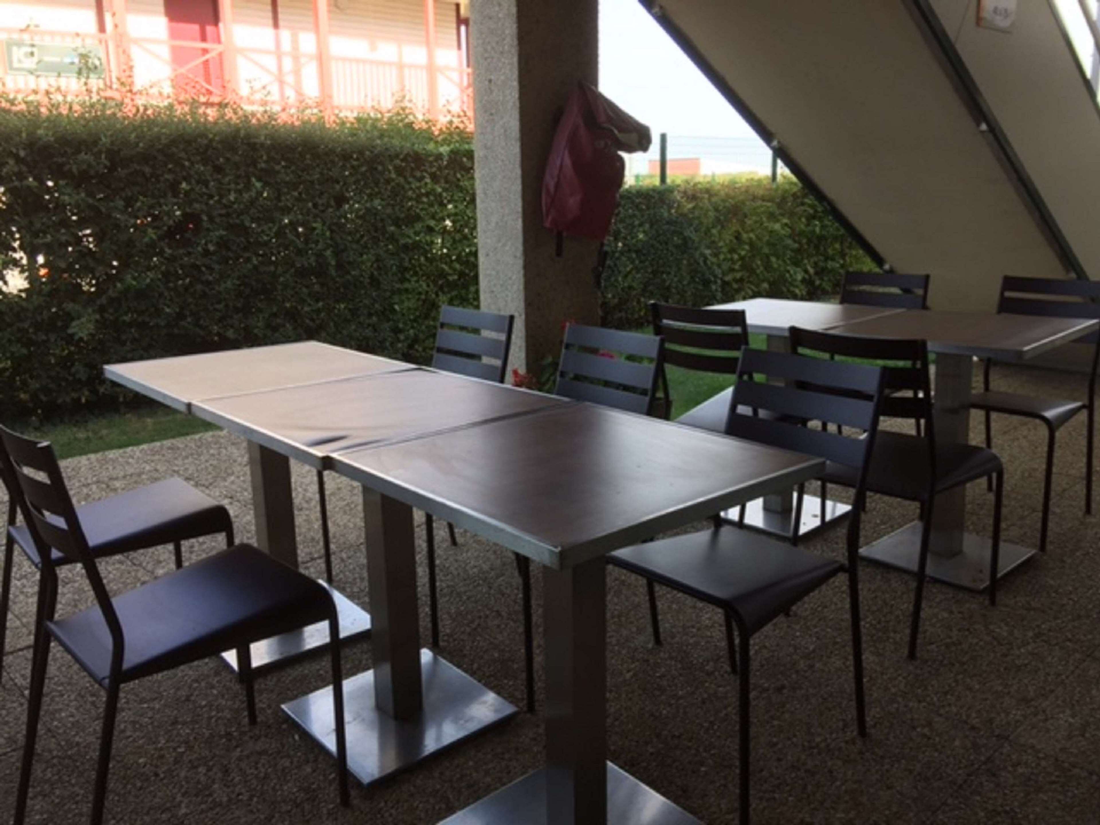 Un área de comedor al aire libre con mesas y sillas, una estructura tipo carpa y un seto al fondo.