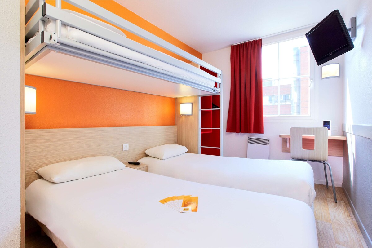 A hotel room with two beds, a desk, a chair, a TV, and a window with red curtains.