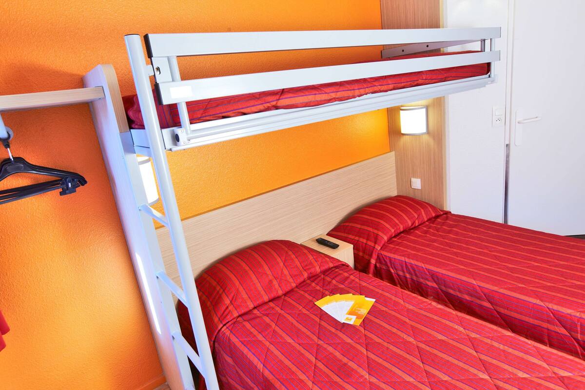 A bunk bed with red bedding, a wooden headboard, a small bedside table with a remote control, and a wall-mounted light.