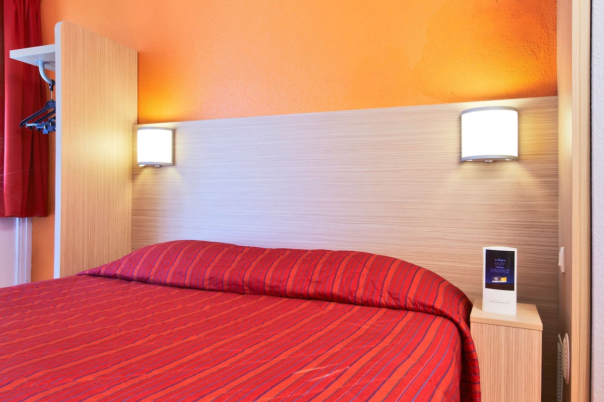 A hotel room with a large bed, red bedding, two wall-mounted lights, and a small bedside table with a sign.