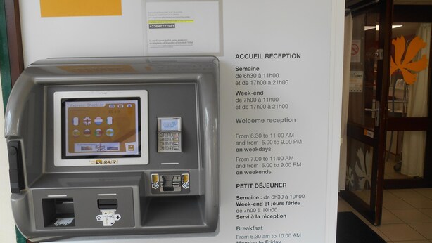 Un mostrador de recepción automático con pantalla digital y lector de tarjetas, un número de contacto disponible las 24 horas del día, los 7 días de la semana, y el horario de atención al cliente indicado.
