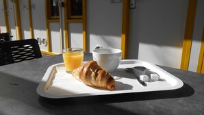 Bandeja de desayuno con un croissant, un vaso de jugo de naranja y un bol sobre una mesa, con una ventana al fondo.