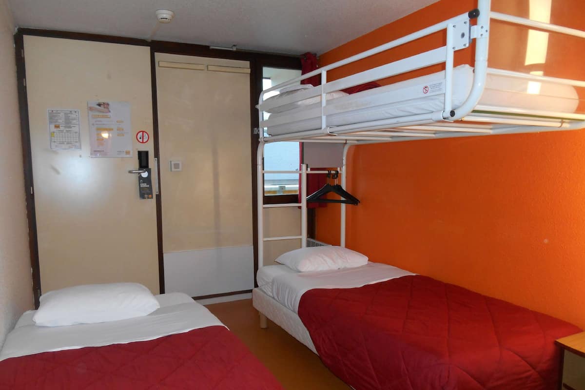 Habitación de residencia con dos camas, una con una manta roja y la otra con una almohada blanca. Hay una cama nido encima de las camas y una puerta con una cámara de seguridad y un aviso.