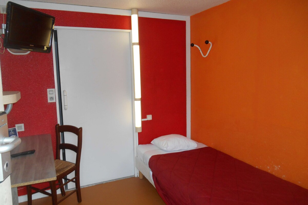 Ein kleines Zimmer mit einem Bett, einem Sessel, einem Schreibtisch und einem Fernseher.
