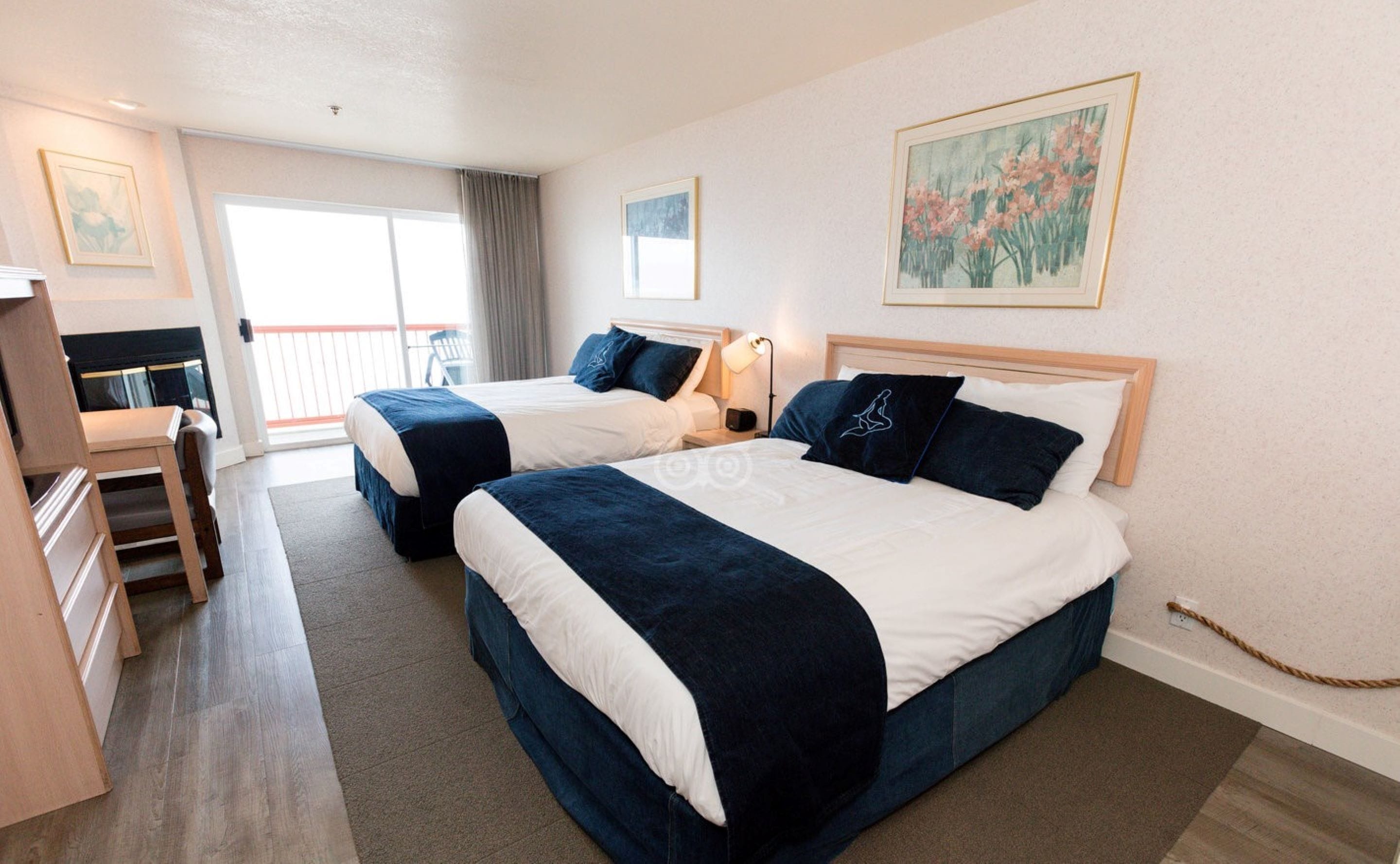 A hotel room with two beds, a desk, and a chair. There is a painting on the wall and a view of the outside balcony.