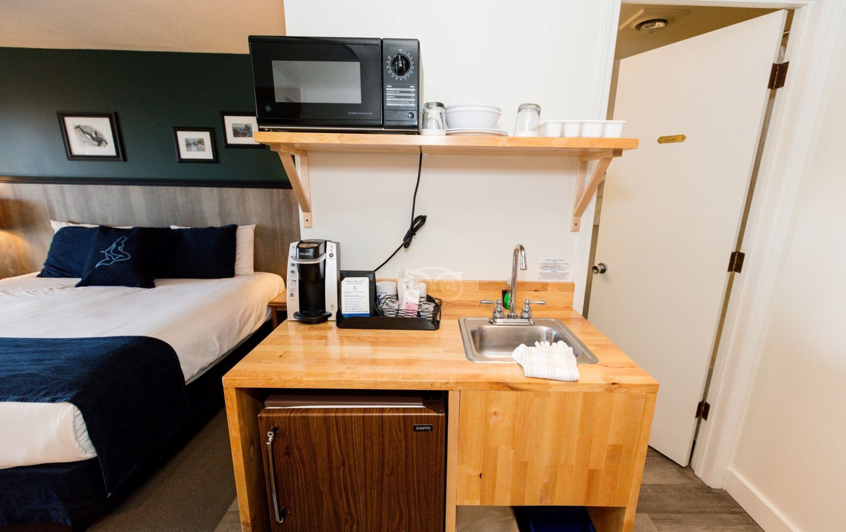 A compact hotel room with a bed, a small kitchenette, a microwave, a coffee machine, and a sink.