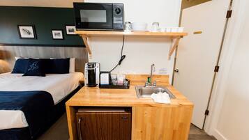 A compact hotel room with a bed, a small kitchenette, a microwave, a coffee machine, and a sink.