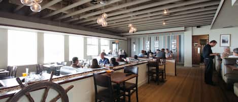 A restaurant with a high ceiling, exposed beams, and pendant lighting. There are multiple dining tables and chairs, and a bar area with a large wheel feature.