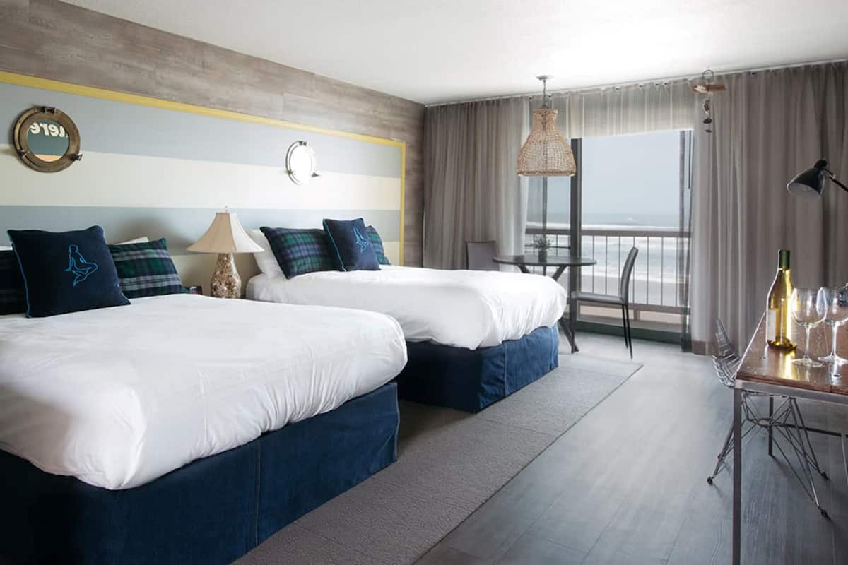 Une chambre d’hôtel avec deux lits, une horloge murale ronde, une lampe, une table avec une bouteille et des verres, et une vue sur l’océan.