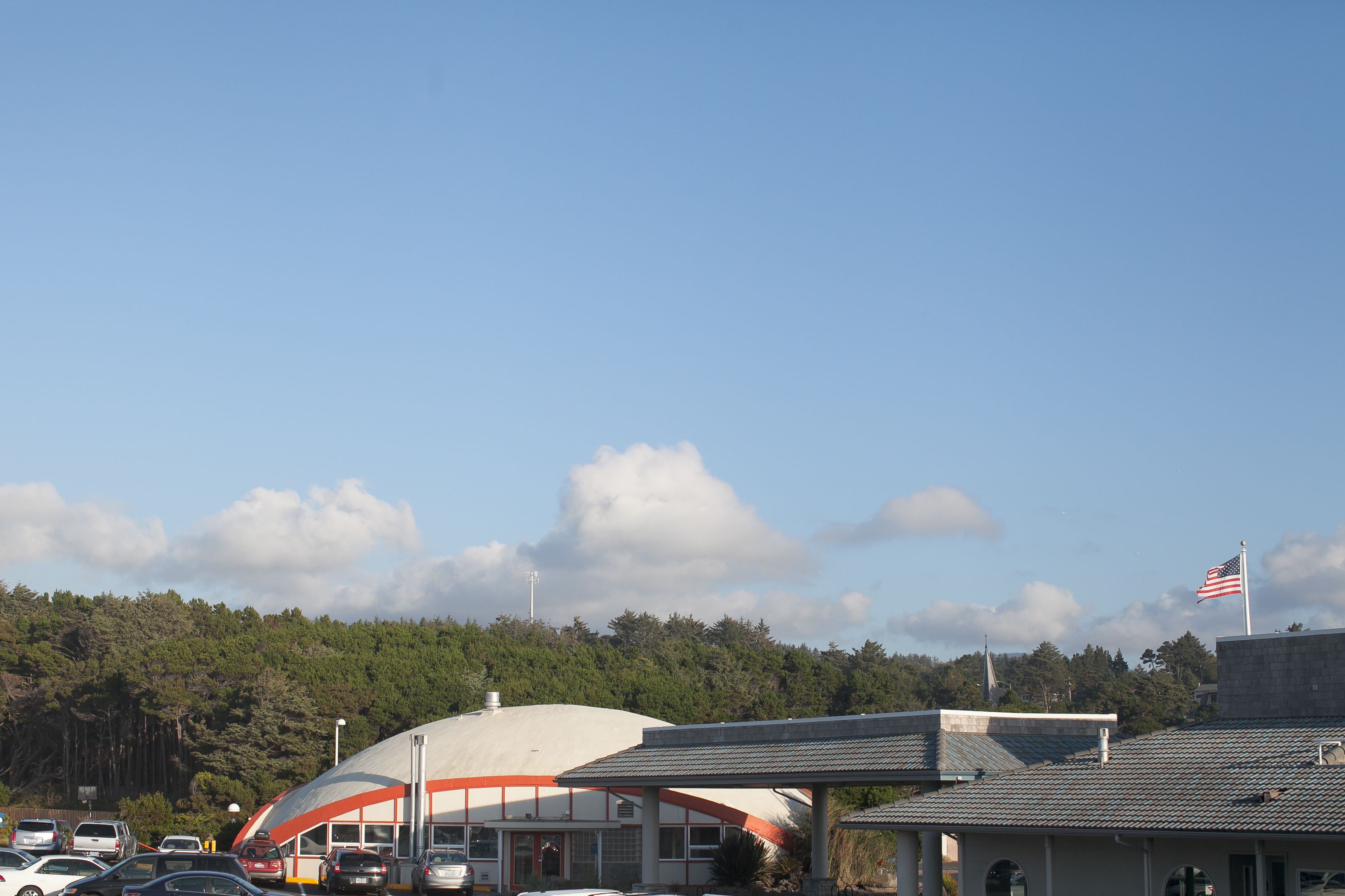 Un parking avec plusieurs voitures, un bâtiment doté d’une structure en forme de dôme et un drapeau sur un mât.