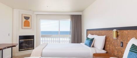 A hotel room with two beds, a fireplace, a view of the ocean, and a balcony.