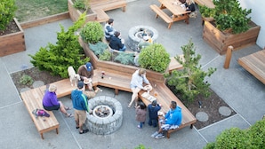 Een terras buiten met houten banken en tafels, omringd door plantenbakken en een vuurkorf in het midden.